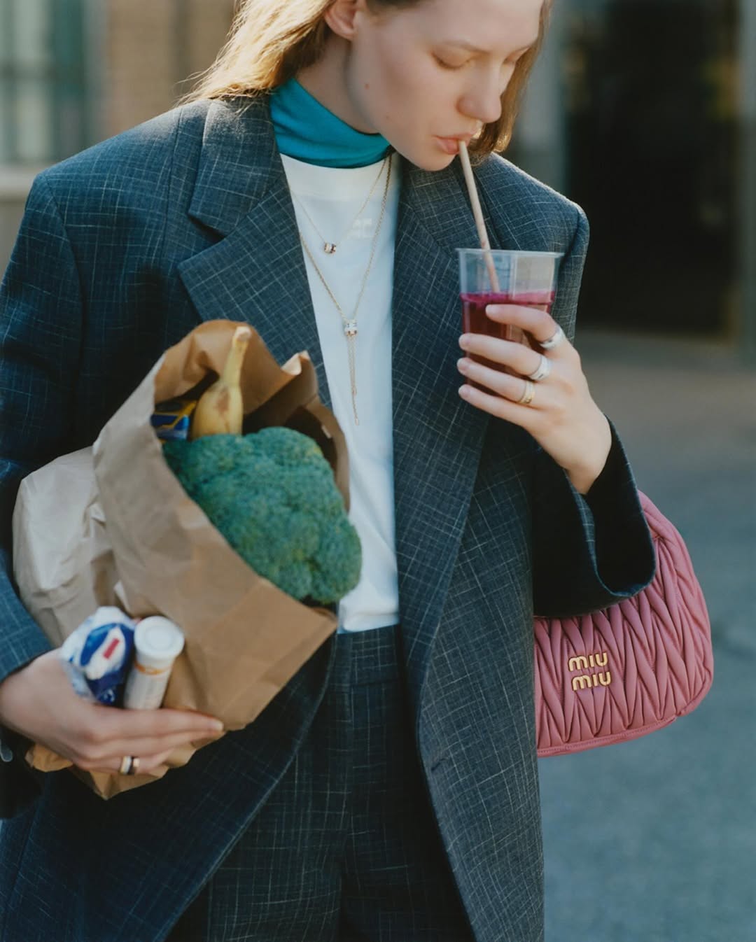 Young woman in grey suit drinks smoothie outdoors while holding grocery bag and pink designer purse, fashion lifestyle shot.