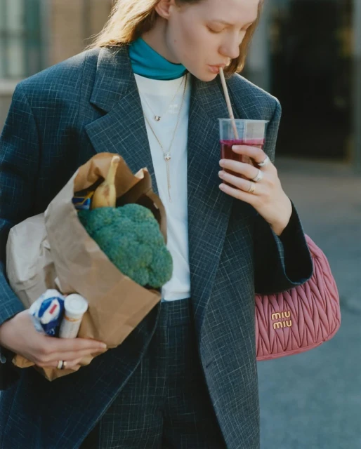 Young woman in grey suit drinks smoothie outdoors while holding grocery bag and pink designer purse, fashion lifestyle shot.