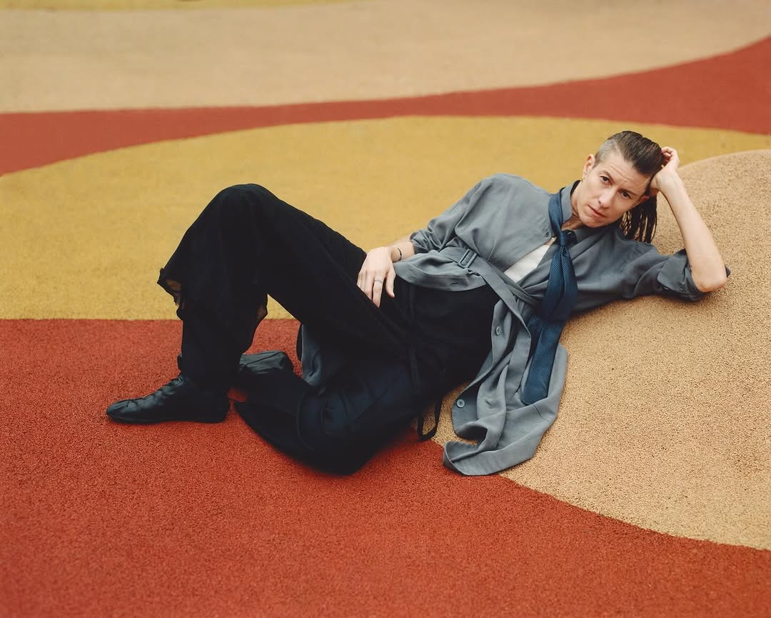 Adult lies on a red and tan surface in a gray shirt and black pants with a blue tie in an outdoor location with medium lighting.