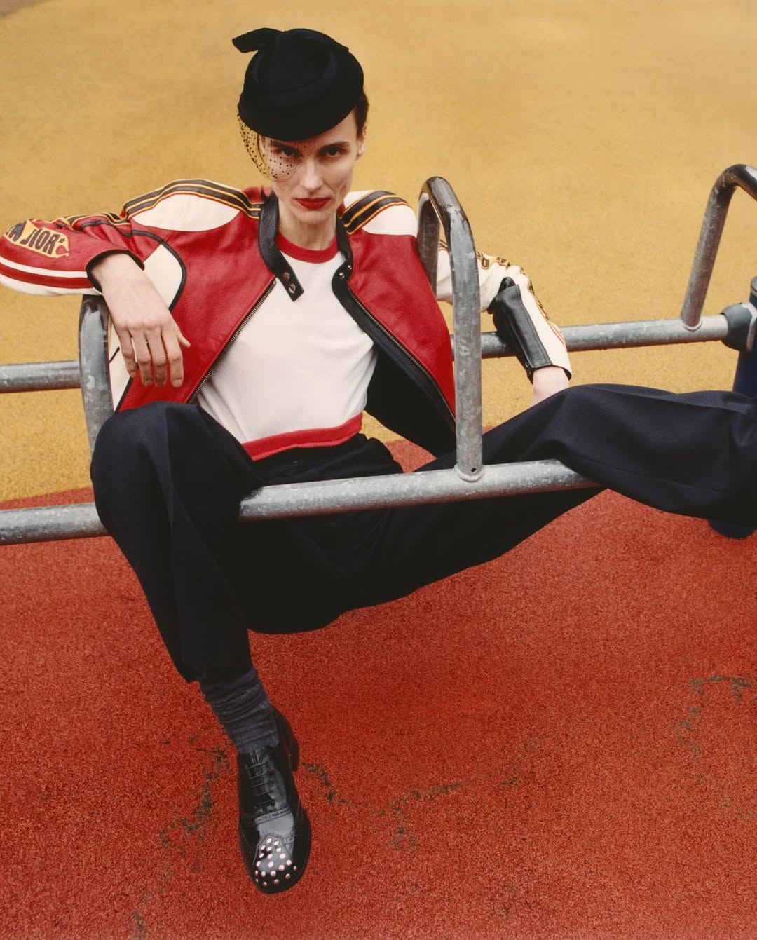 A person sitting on playground equipment wearing a red and white racing jacket, black pants, and a black hat with netting. 
