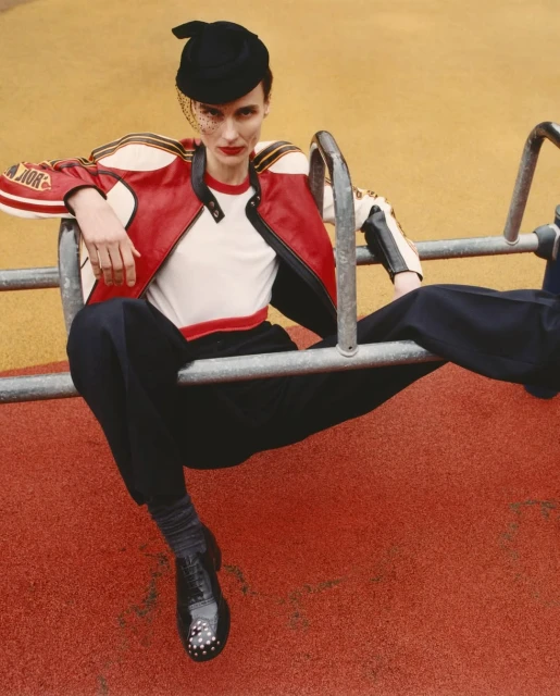 A person sitting on playground equipment wearing a red and white racing jacket, black pants, and a black hat with netting. 