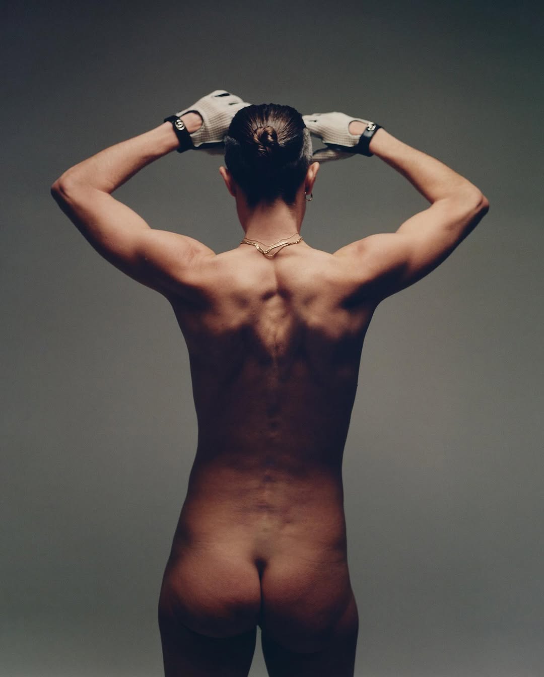 Back view of a muscular, bare-chested adult with hands on their head, wearing gloves, against a neutral studio backdrop.