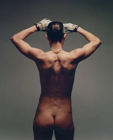 Back view of a muscular, bare-chested adult with hands on their head, wearing gloves, against a neutral studio backdrop.