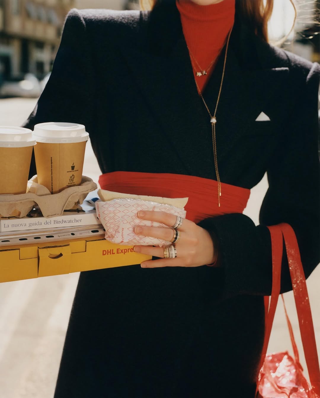 Woman in black coat carries coffee, sandwiches, and a DHL package, showcasing street style and fast delivery against an urban backdrop.