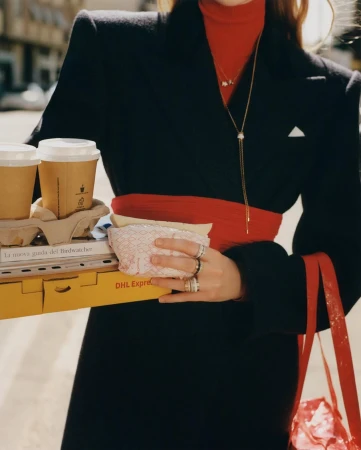 Woman in black coat carries coffee, sandwiches, and a DHL package, showcasing street style and fast delivery against an urban backdrop.