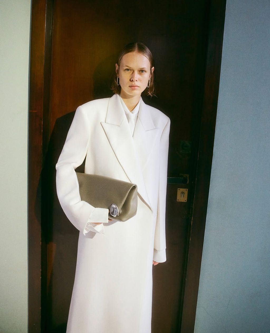 A fair-skinned adult woman poses indoors, holding a light green clutch purse while wearing a long white skirt suit with gold earrings.
