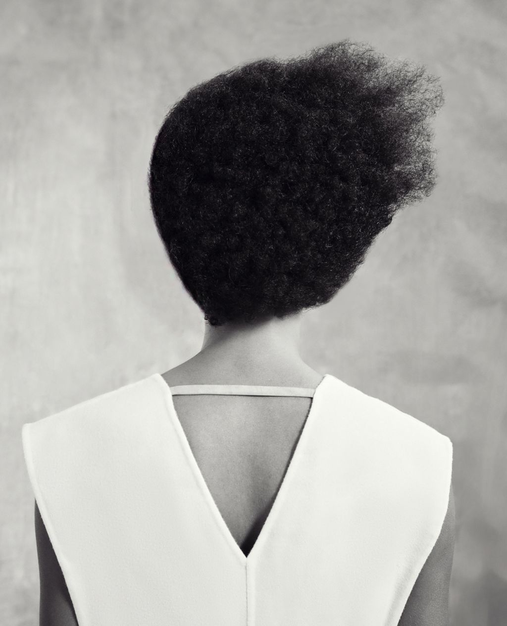 Back view of a woman with dark afro hair wearing a white V-neck top against a mottled grey background in a studio shot.