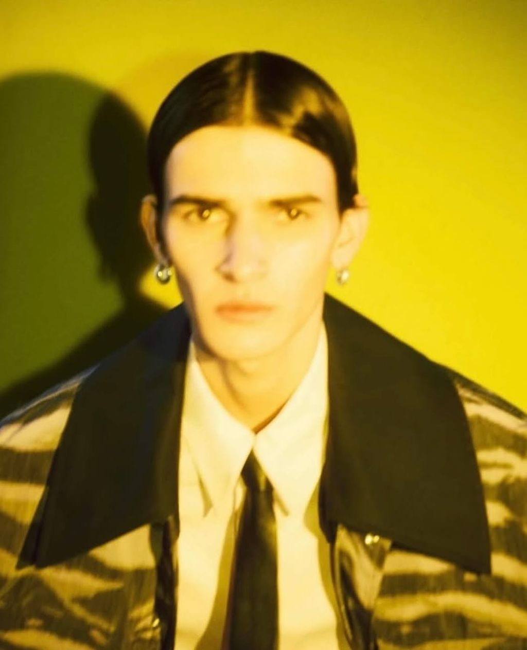 Headshot of a slender adult male with dark hair, light skin, and an expressionless face wearing a white shirt, black tie, and jacket against a yellow backdrop.
