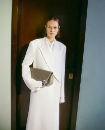 Model in a structured white coat holding a grey clutch bag standing in a doorway by Armando Cherillo