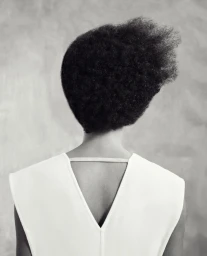 Rear view of a person with short textured black hair wearing a white top with a v-neck back against a neutral textured background.