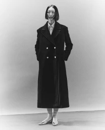Model in a long black double-breasted coat standing against a light grey studio background by Armando Cherillo