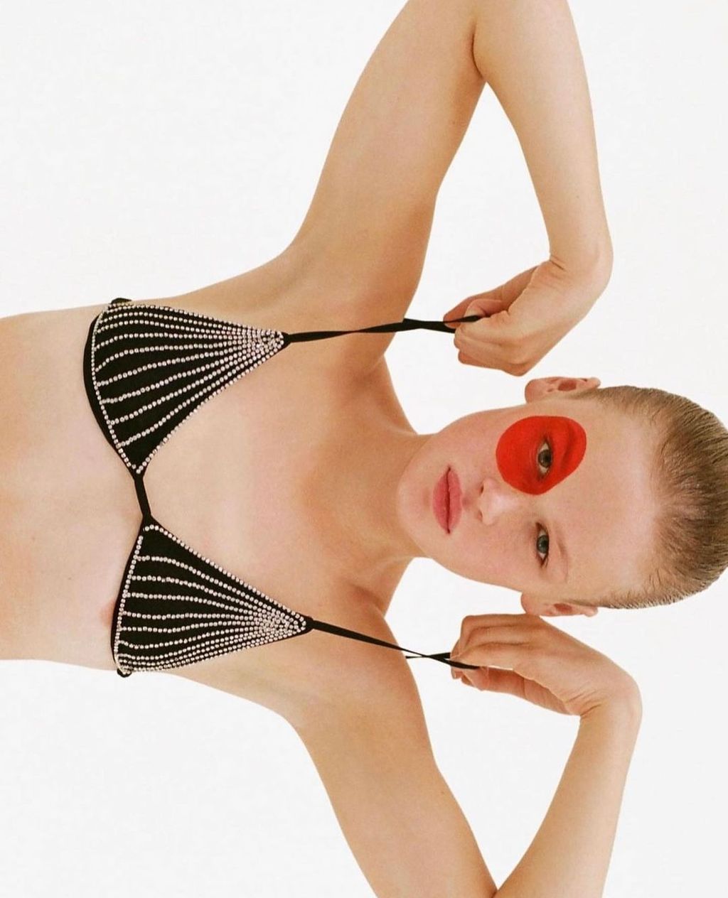 Attractive young woman poses in bikini top, one eye covered with a red circle, on a white backdrop for a beauty and fashion shot.