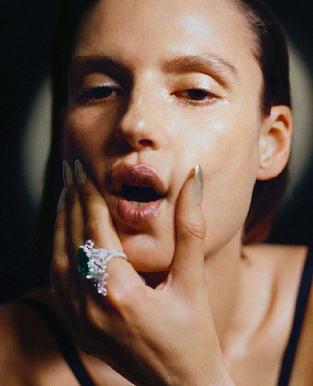 Close-up studio portrait of a fair-skinned adult female with silver nails and a large gemstone ring, holding her face in her hand.