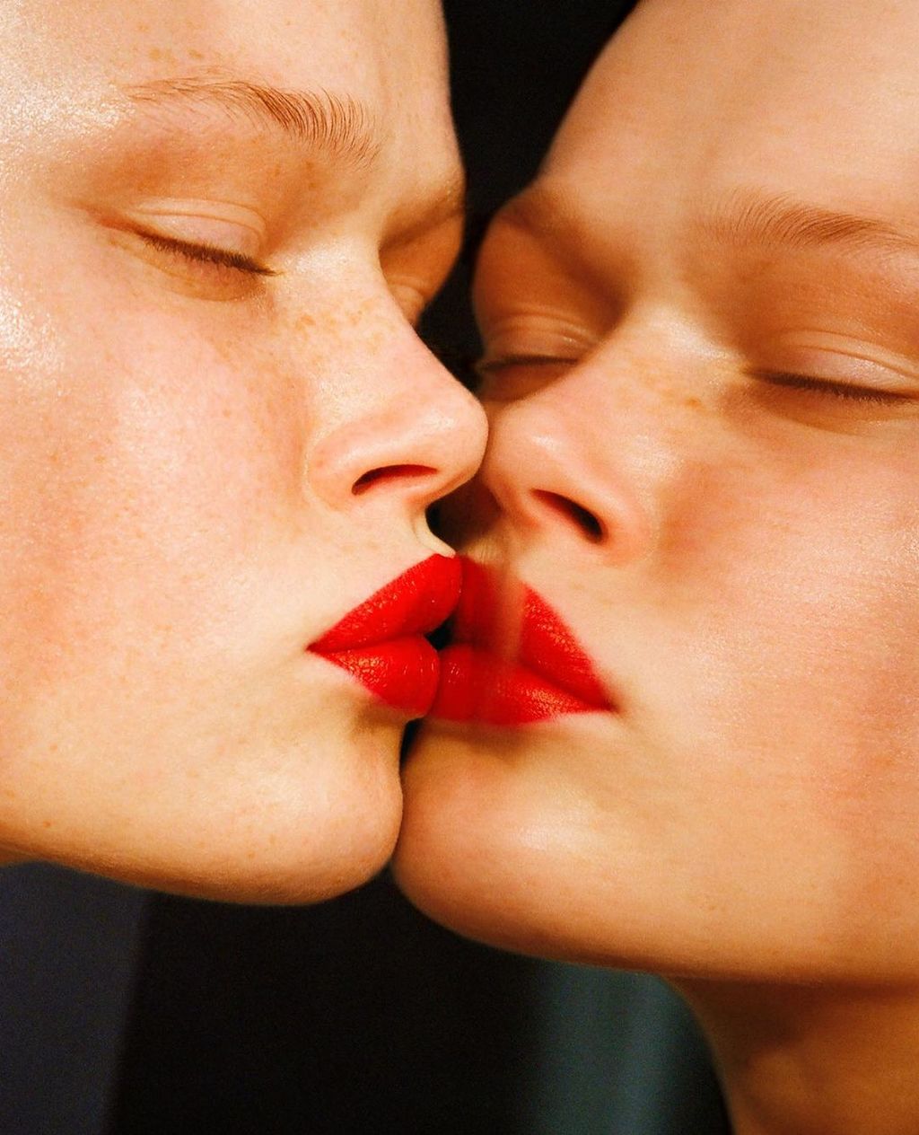 Close-up of two fair-skinned adult females kissing with bright red lipstick, intimate skin contact, eyes closed, dark background.