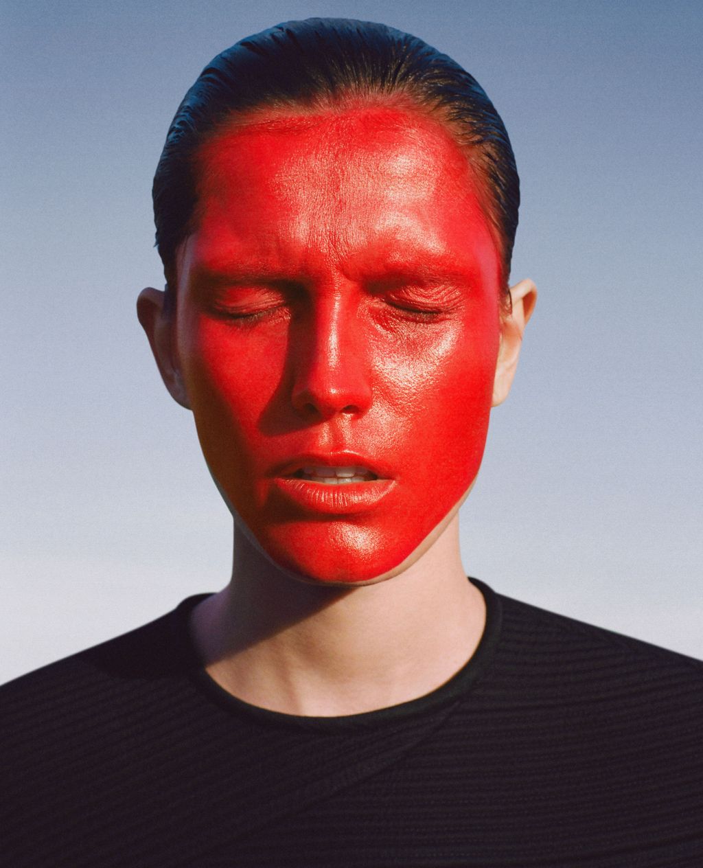 Close-up portrait of an adult female with red face paint, eyes closed, wearing a dark shirt against a light blue sky backdrop.