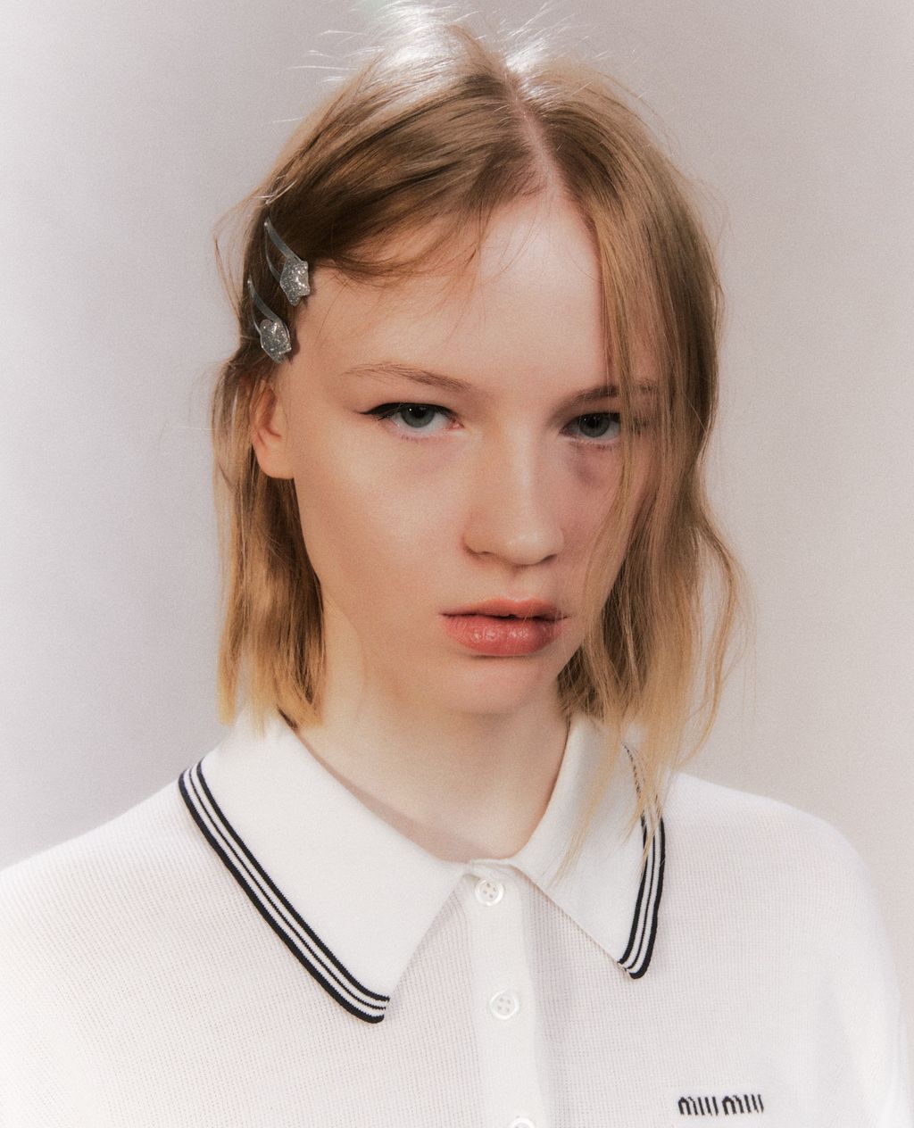 Close-up studio shot of a blond woman with hair clips, wearing a white collared shirt with black trim, and the brand logo at the hem.