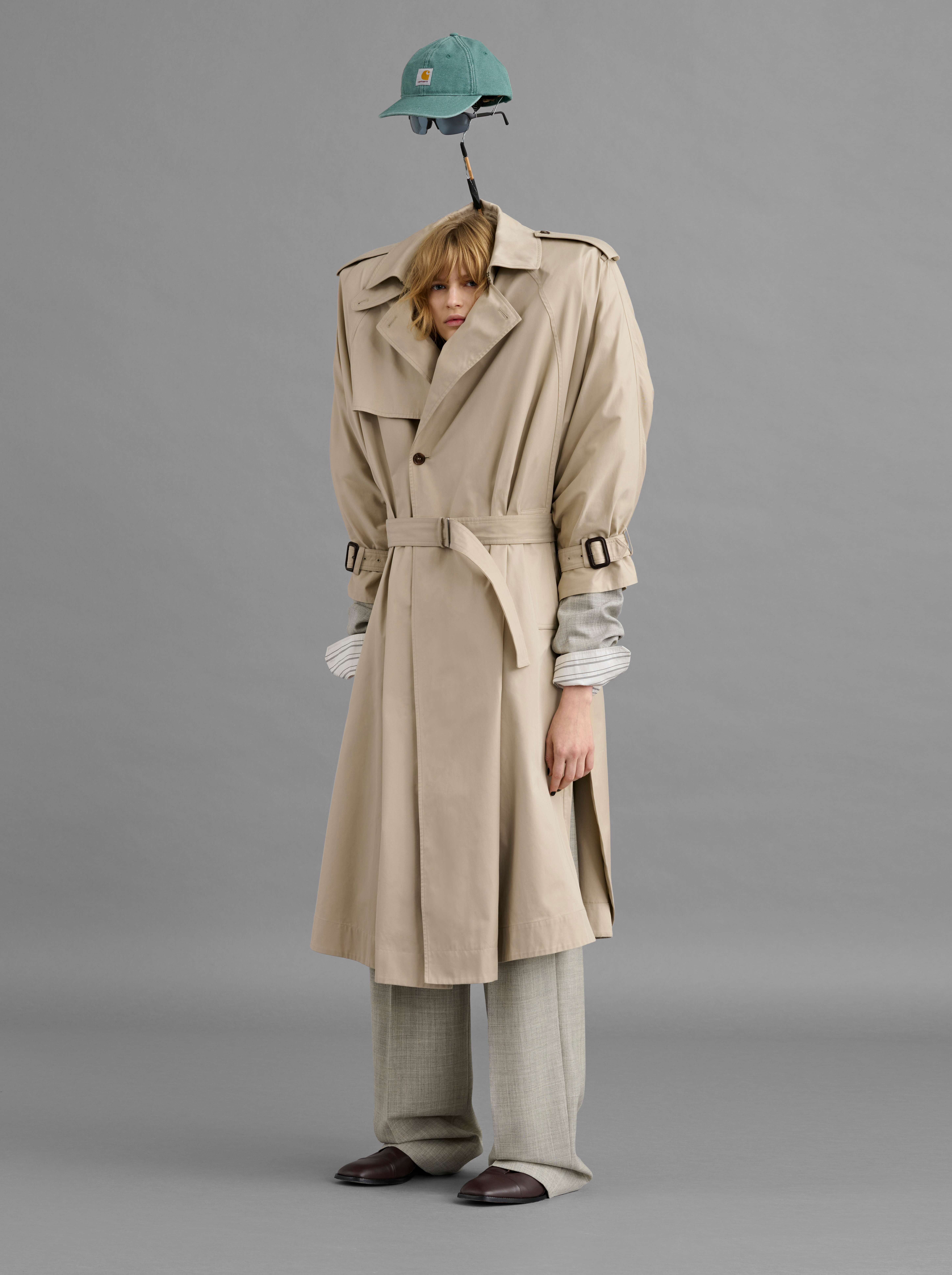 Fashionable woman in a tan trench coat and grey trousers poses in a studio, wearing a baseball cap and sunglasses on her head.