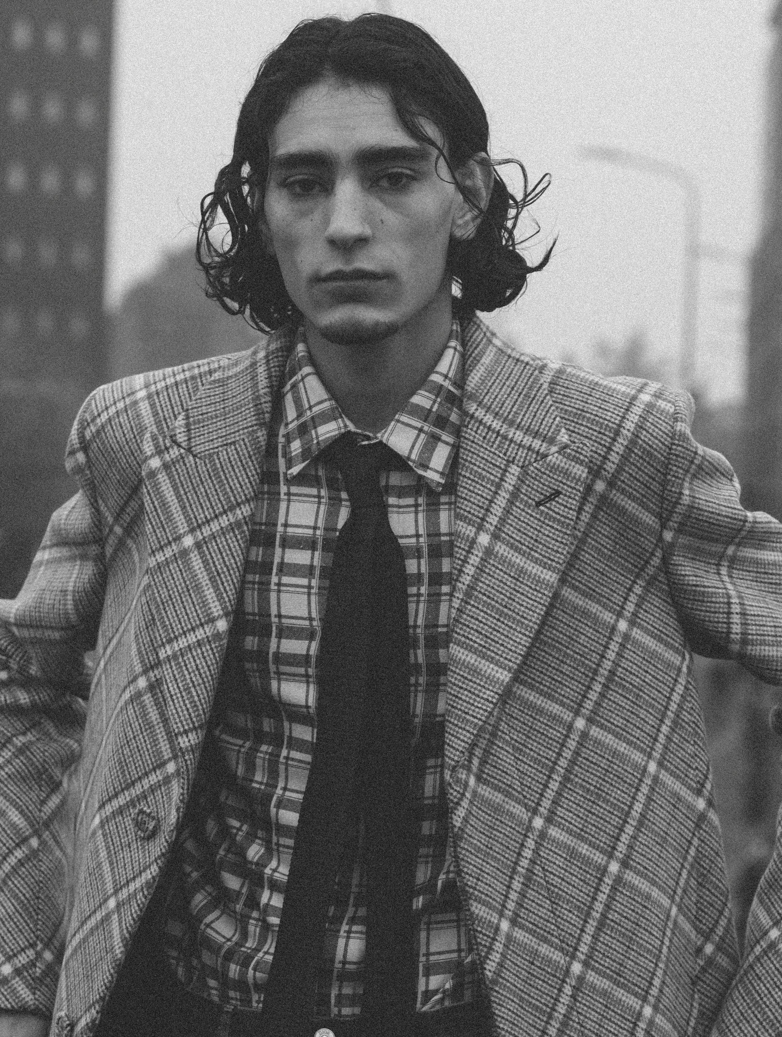Monochrome portrait of a young adult male wearing a plaid suit and black tie standing outdoors with buildings in the distance.