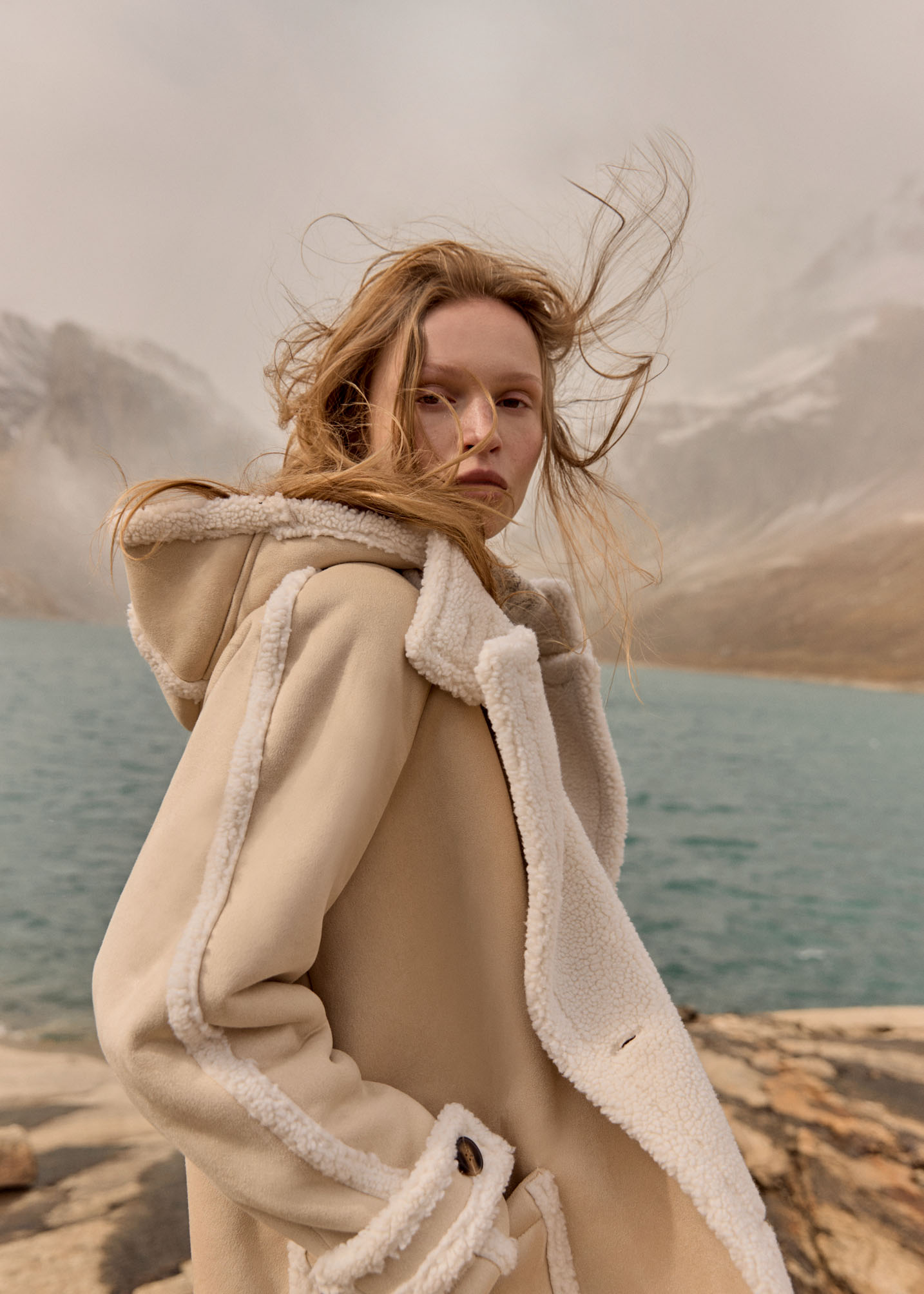 Woman in a tan hooded coat with a faux fur trim stands in a mountainous landscape with a turquoise lake under an overcast sky.