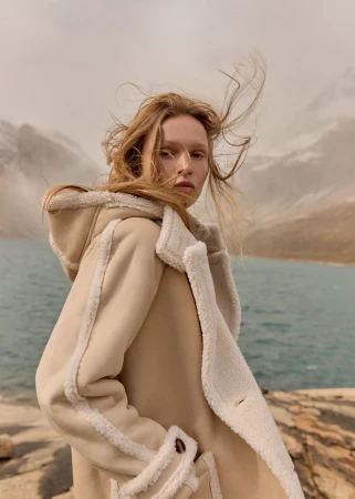 Woman in a tan hooded coat with a faux fur trim stands in a mountainous landscape with a turquoise lake under an overcast sky.