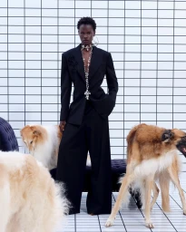 Model in a black suit standing in front of a white tiled wall with long-haired dogs in the foreground