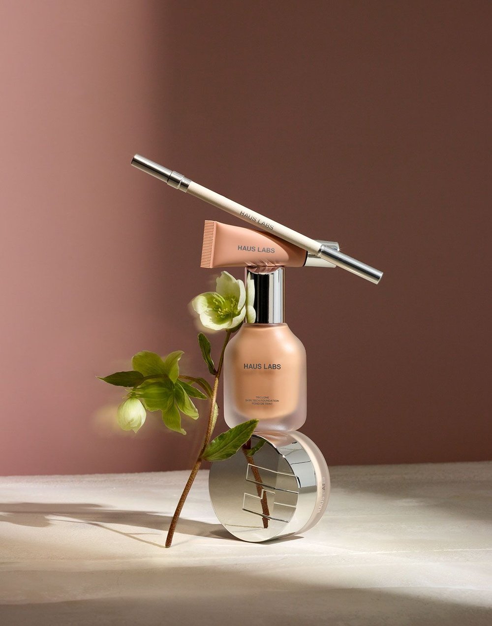 Haus Labs makeup products displayed on a table with a hellebore flower against a mauve background in a beauty product shot.