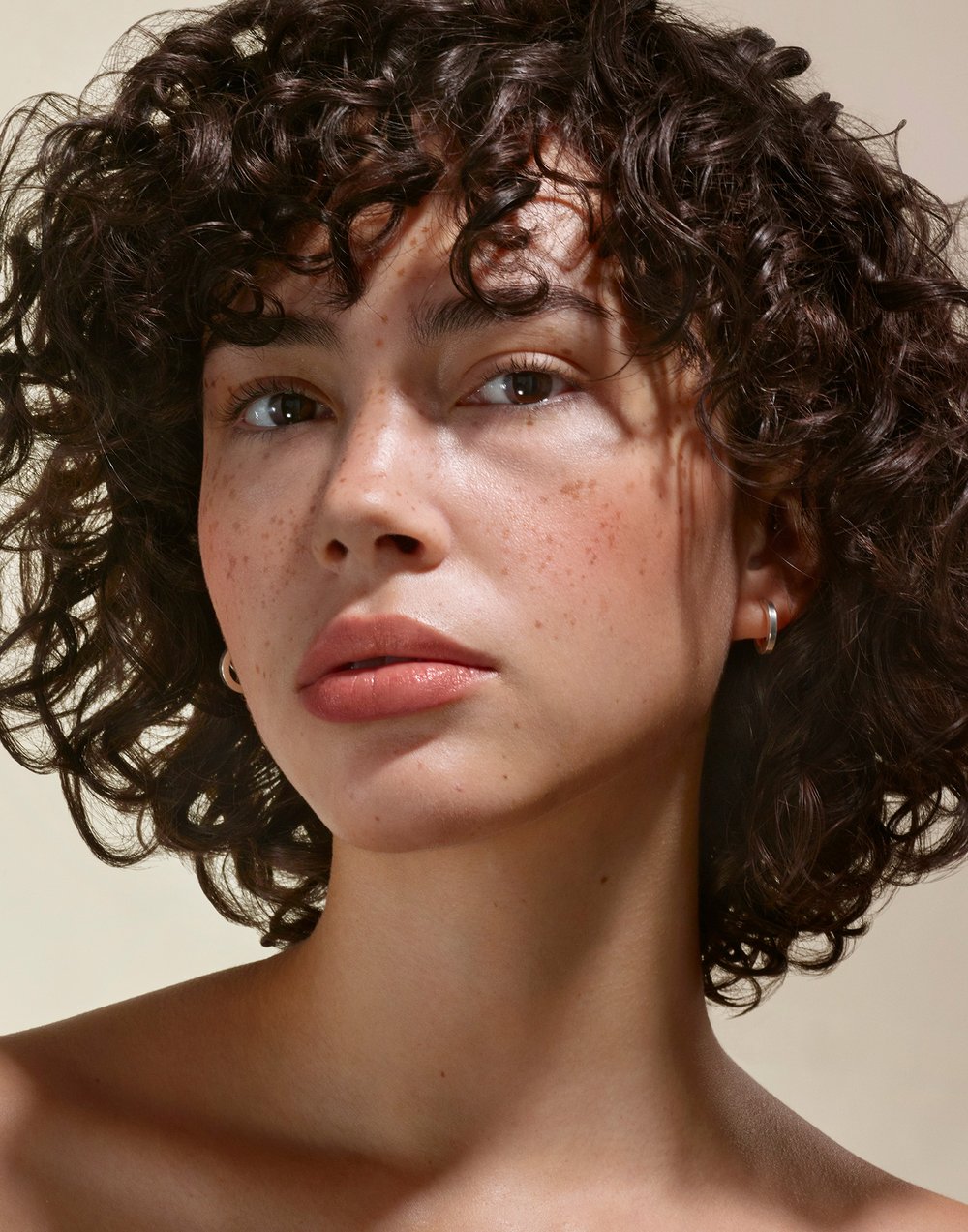 Close-up portrait of a woman with curly brown hair and freckles, wearing hoop earrings, with natural makeup, against a neutral background.