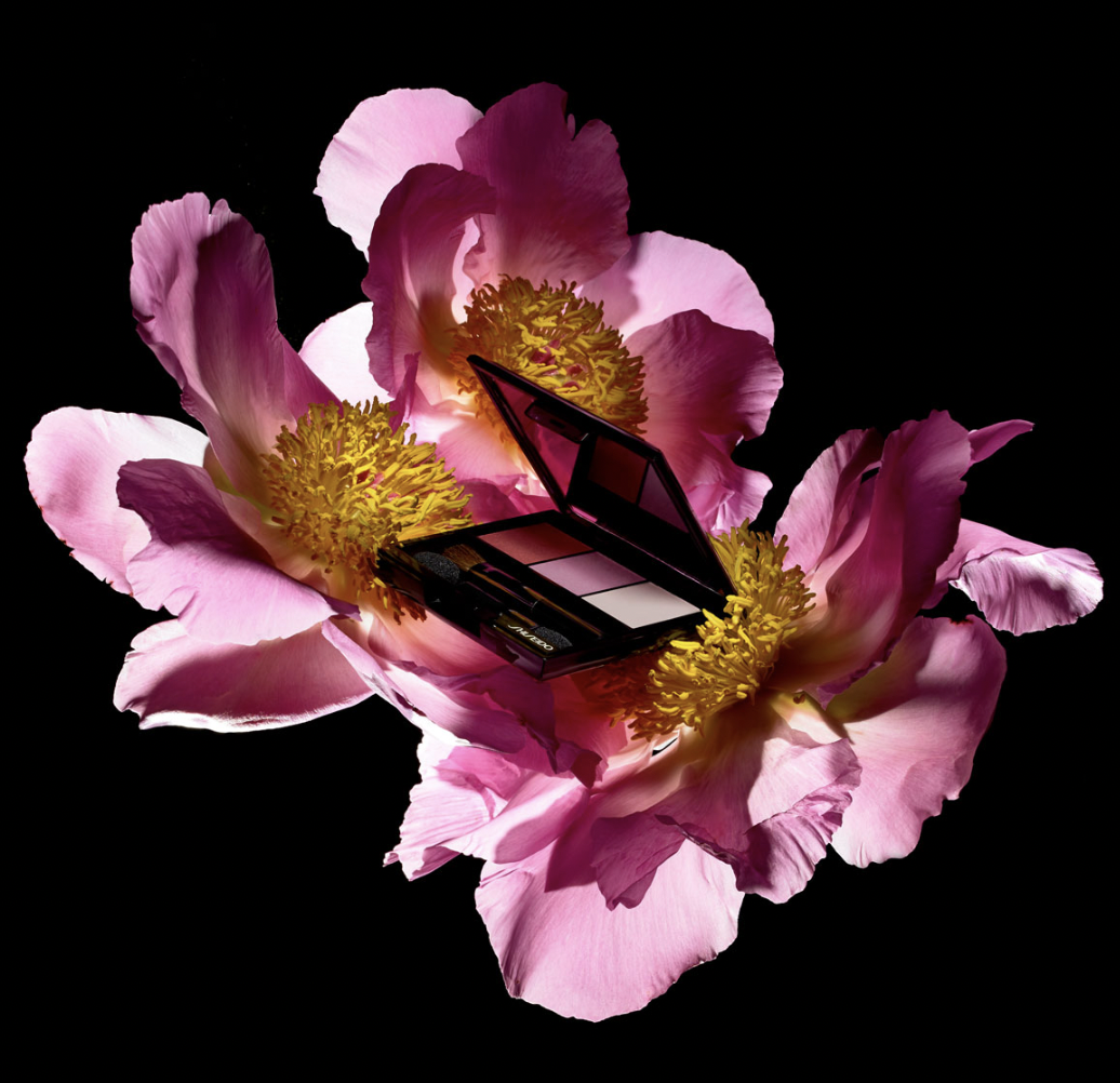 Open cosmetic compact on pink peony flowers against a black backdrop, with a small brush included for beauty or fashion advertising.