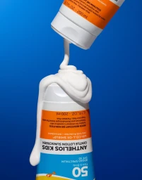 White sunscreen lotion pouring from an inverted tube onto another sunscreen bottle against a vivid blue background