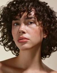 Close-up portrait of a woman with dark curly hair and freckles looking directly at the camera against a neutral background.