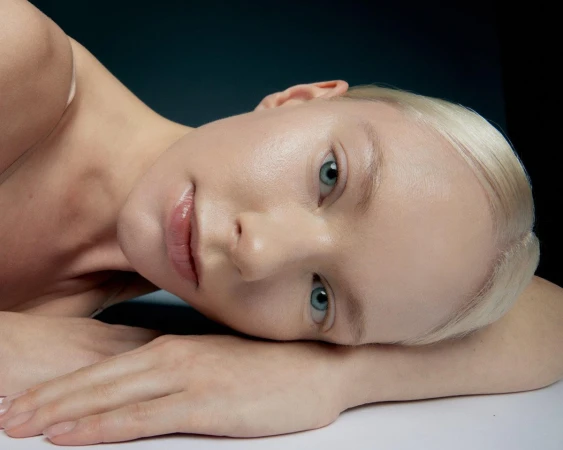 Close-up studio portrait of a fair-skinned adult female model with blue eyes, short blonde hair, glossy lips, and hands near her face.