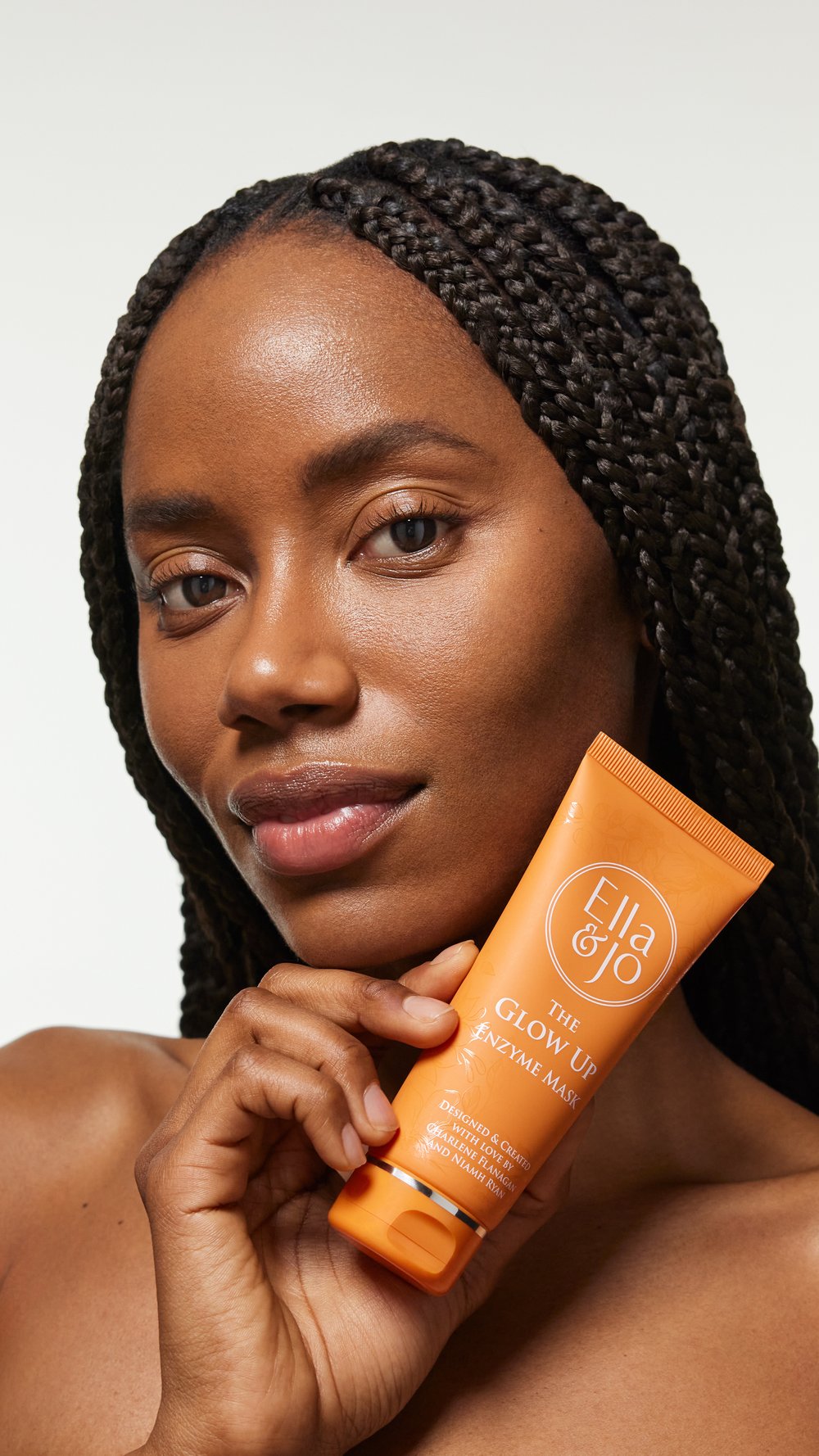 Close-up of a smiling African-American woman holding a tube of Ella & Jo The Glow Up Enzyme Mask against a plain light background.