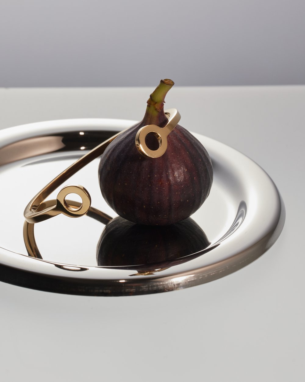 Still life shot of a golden bracelet on a silver tray with a fig. The bracelet has a geometric design with circular elements.