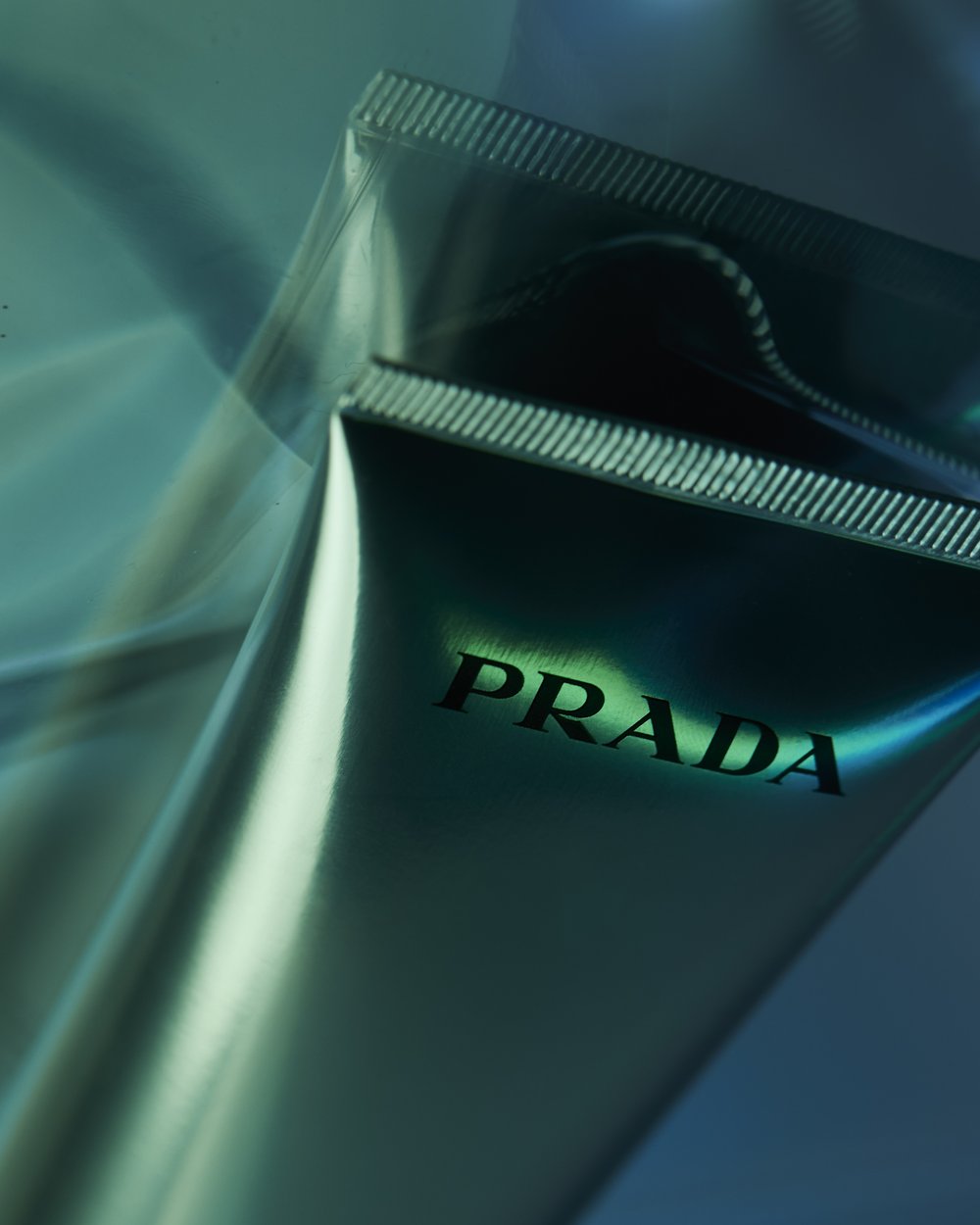 Close-up of Prada branded product packaging with a metallic sheen against a blue and turquoise gradient backdrop in soft studio lighting.