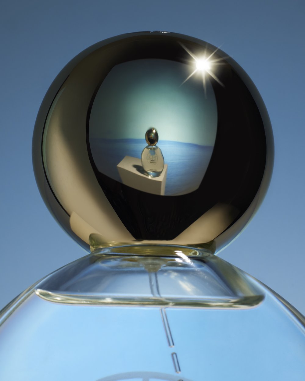 Still life shot of a perfume bottle in a glass orb reflecting a serene seascape under a clear blue sky, evoking feelings of calm.