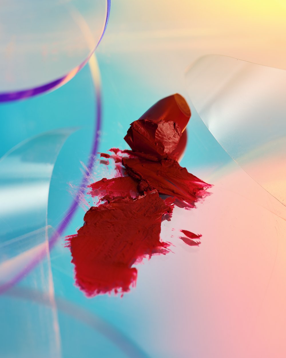 Striking close-up of a broken red lipstick on a reflective surface with vibrant blue and yellow gradients, creating a modern beauty shot.