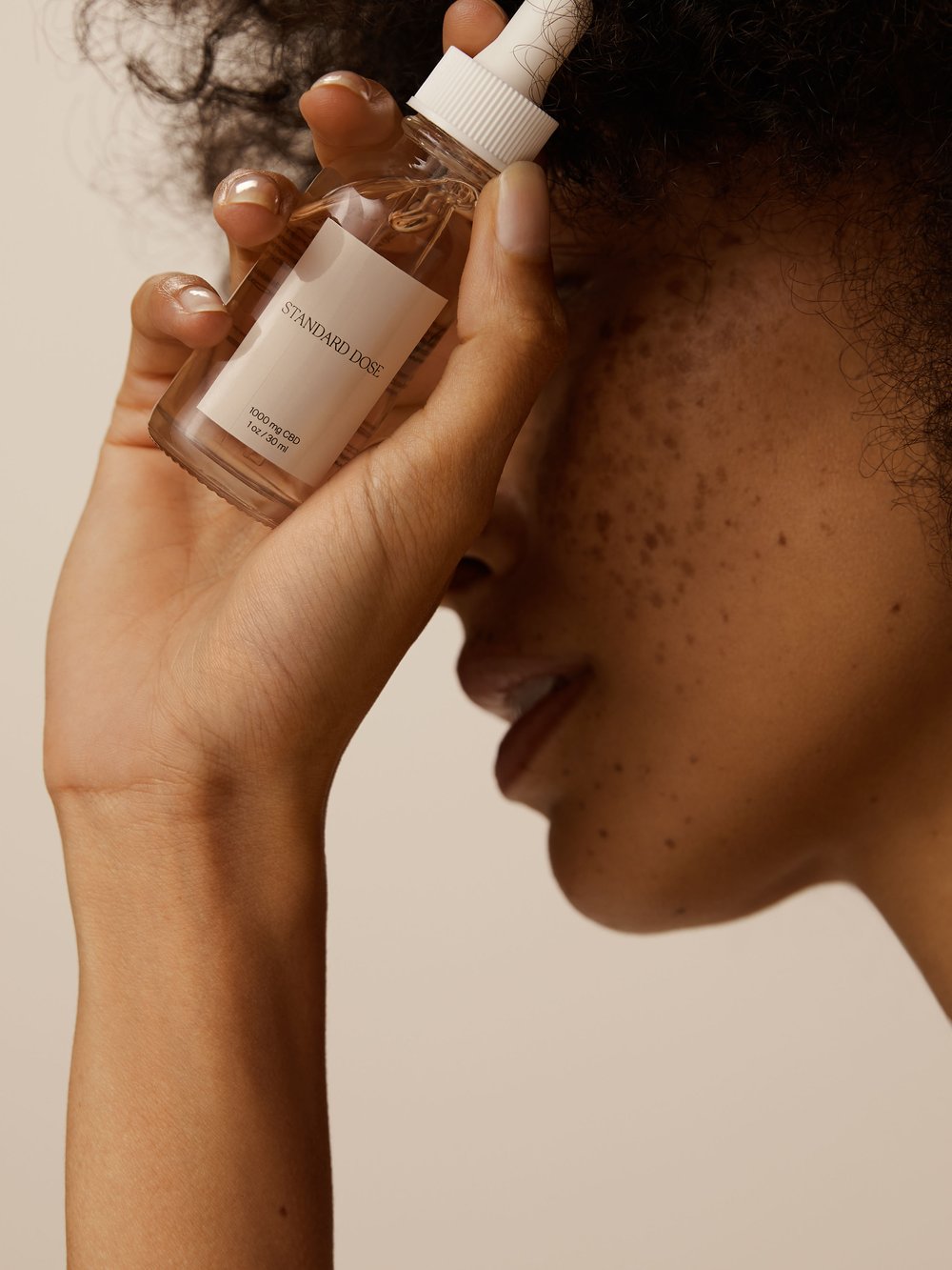 Close up of a fair-skinned adult female with freckles holding a bottle of serum with a dropper against a neutral beige backdrop.