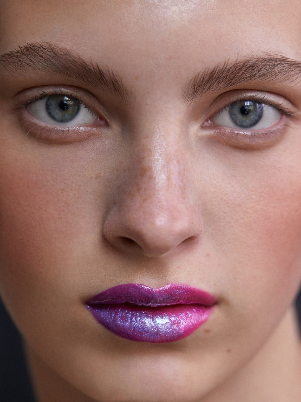 Close-up portrait of a young woman with blue eyes and pink and purple iridescent lipstick, showcasing makeup artistry and beauty.