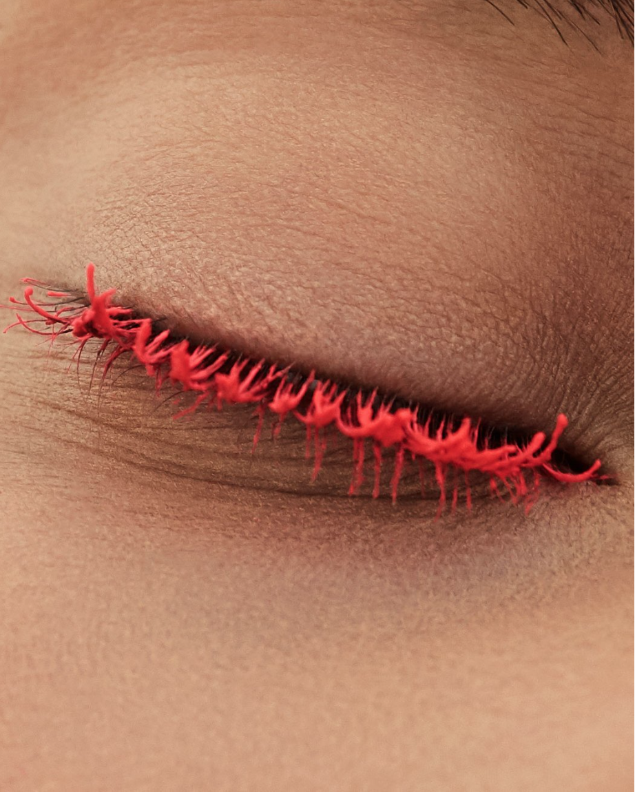 Close-up of a closed eye with bright red, stylized eyelashes. A macro shot highlights the texture of the skin.