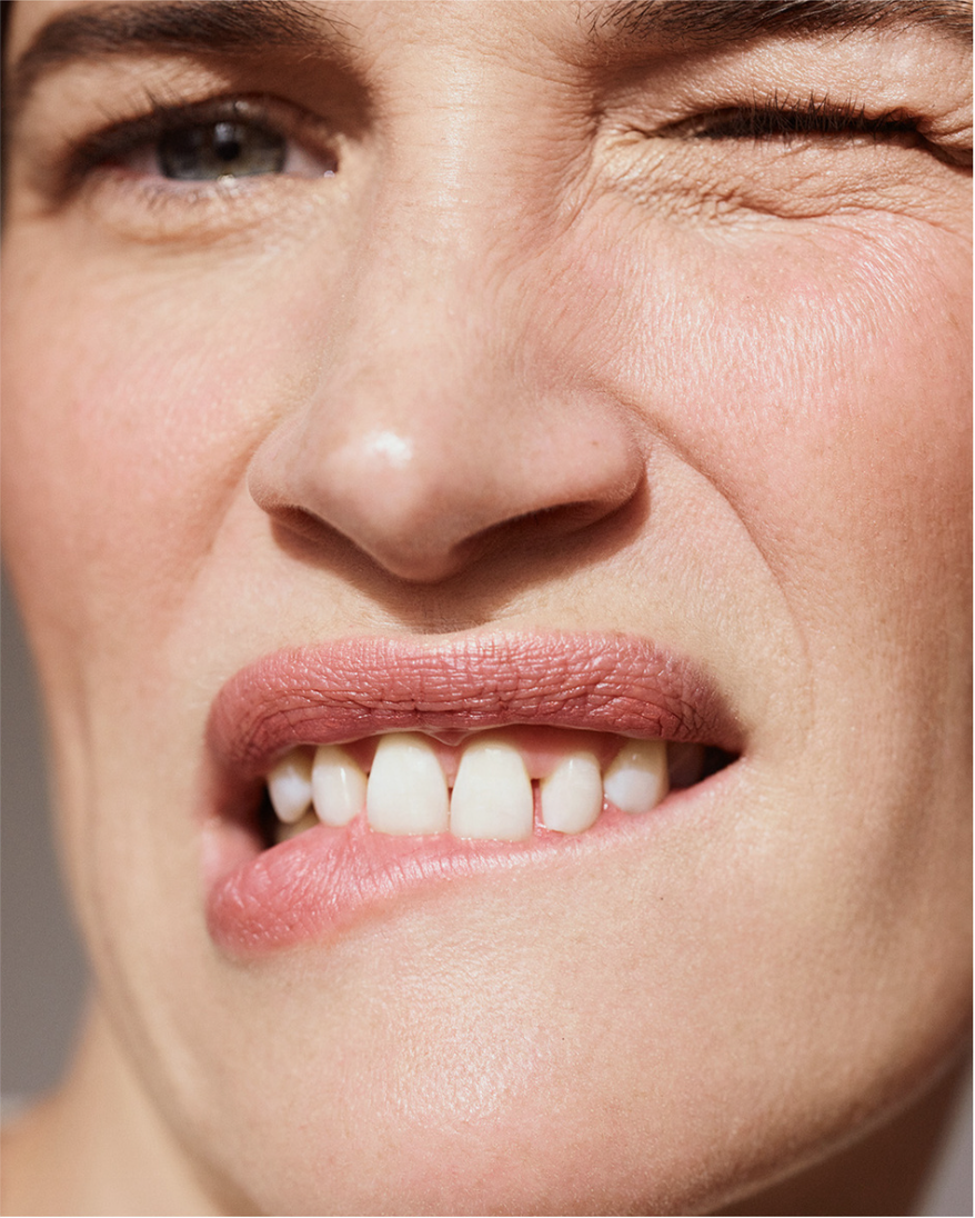 Close-up studio portrait of a fair-skinned adult woman with a winking expression and biting her lip, highlighting facial features and skin texture.