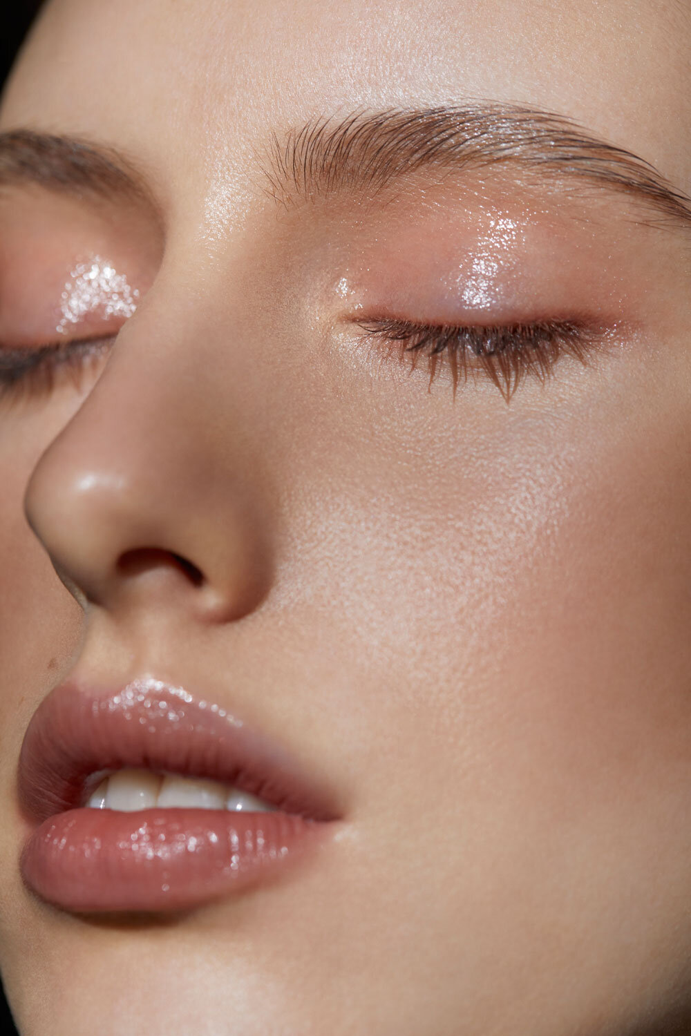 Close-up studio shot of a fair-skinned young adult female face with glossy neutral makeup and closed eyes against a dark background.