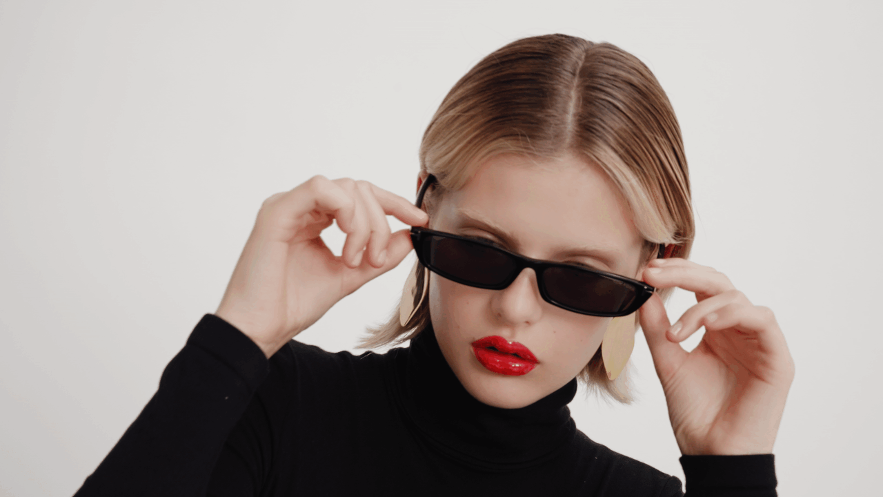 A fair-skinned young woman with glossy red lips poses in a black turtleneck and sunglasses against a stark white background.