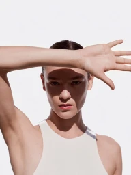 Close-up portrait of a woman wearing a white tank top with her arm raised across her forehead casting a shadow on her face against a white background.