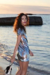 Woman in a sparkling silver mini dress walking near the ocean at sunset, captured in motion with soft blur.