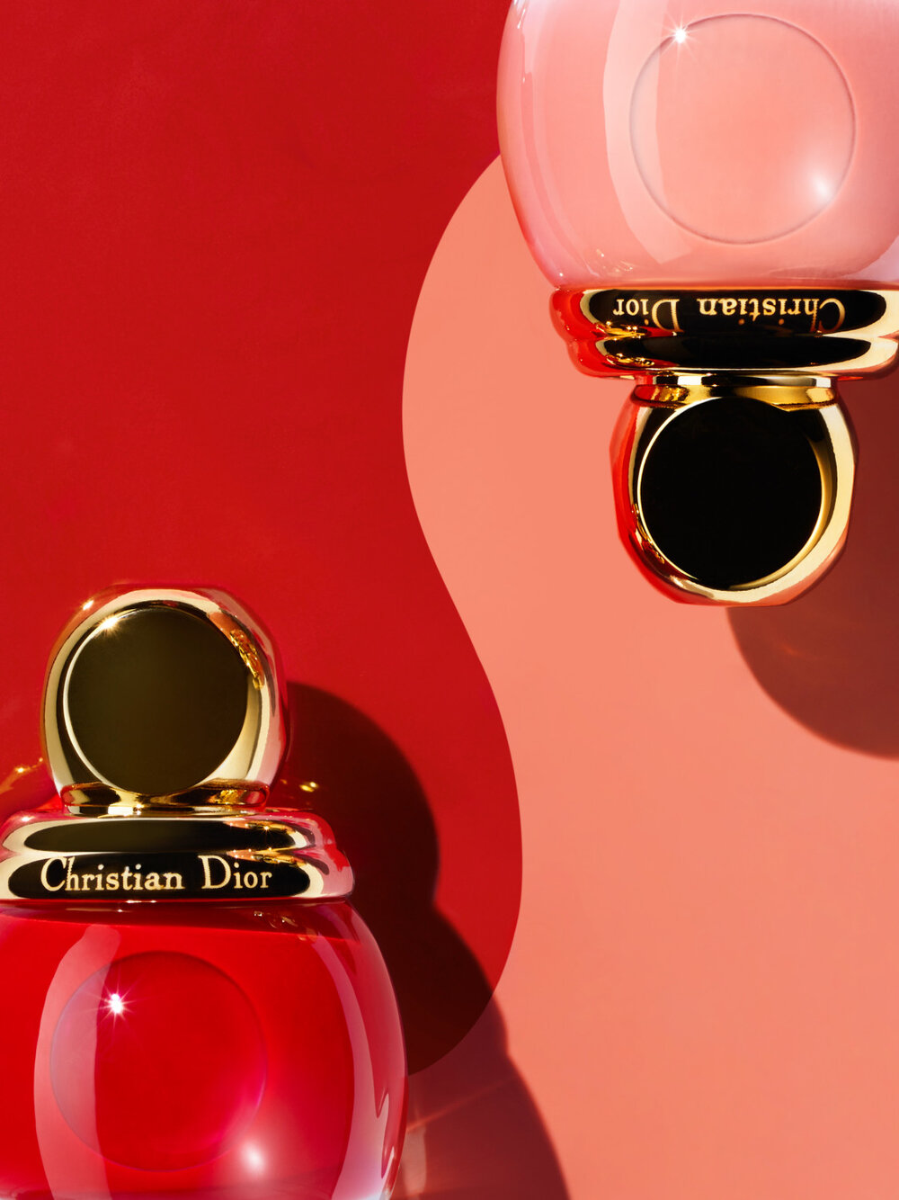 Still life close-up studio shot of red and pink Dior nail polish bottles with gold caps on a red and pink curved backdrop.