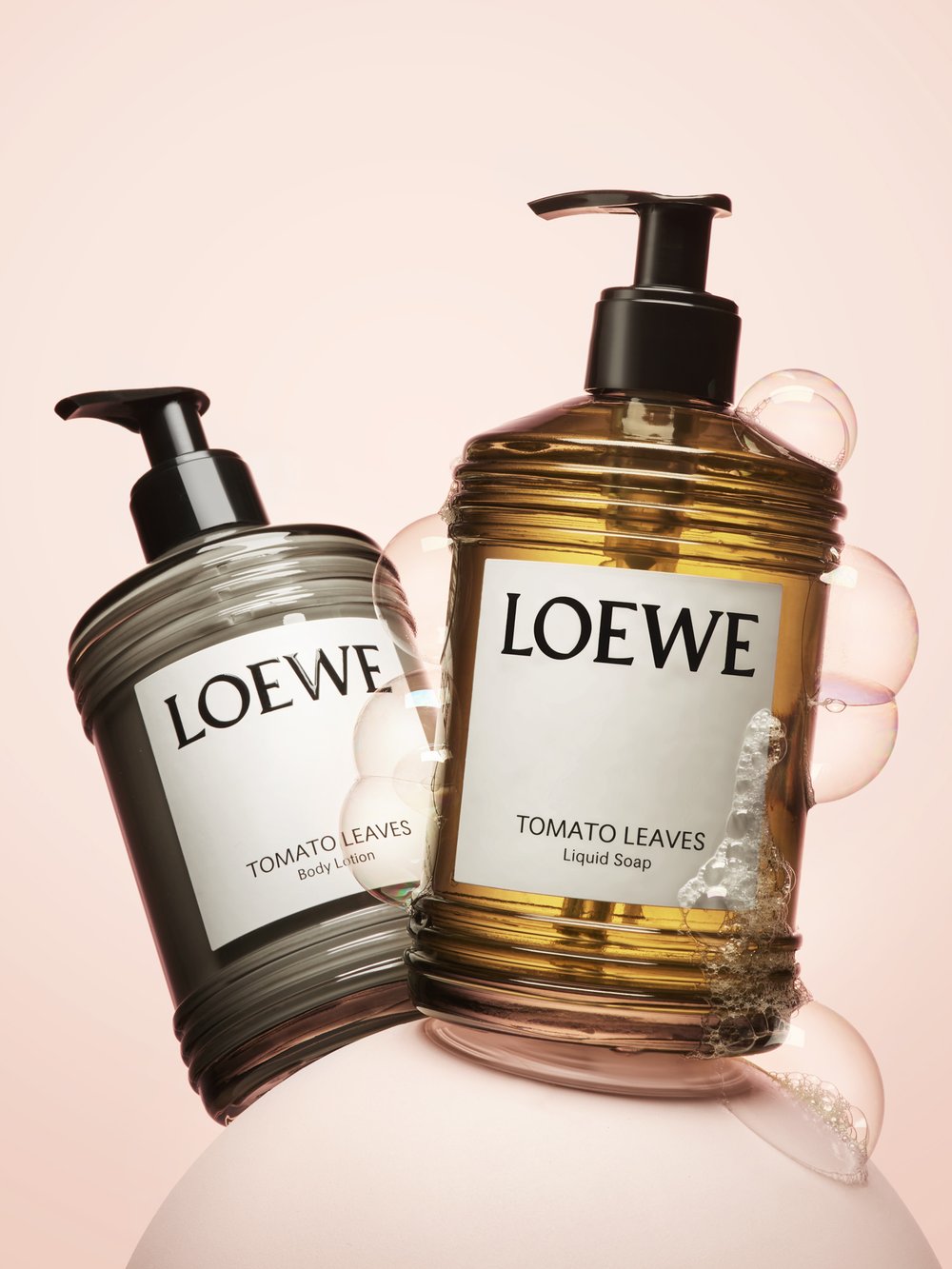 Two bottles of Loewe Tomato Leaves body lotion and liquid soap sit against a pink background, surrounded by soap bubbles.