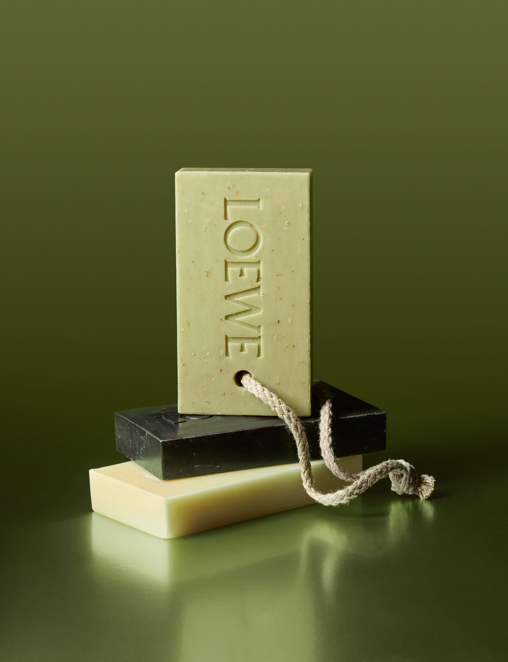 Three bars of soap stacked on top of each other in an indoor setting with a green background. The top bar has LOEWE imprinted.