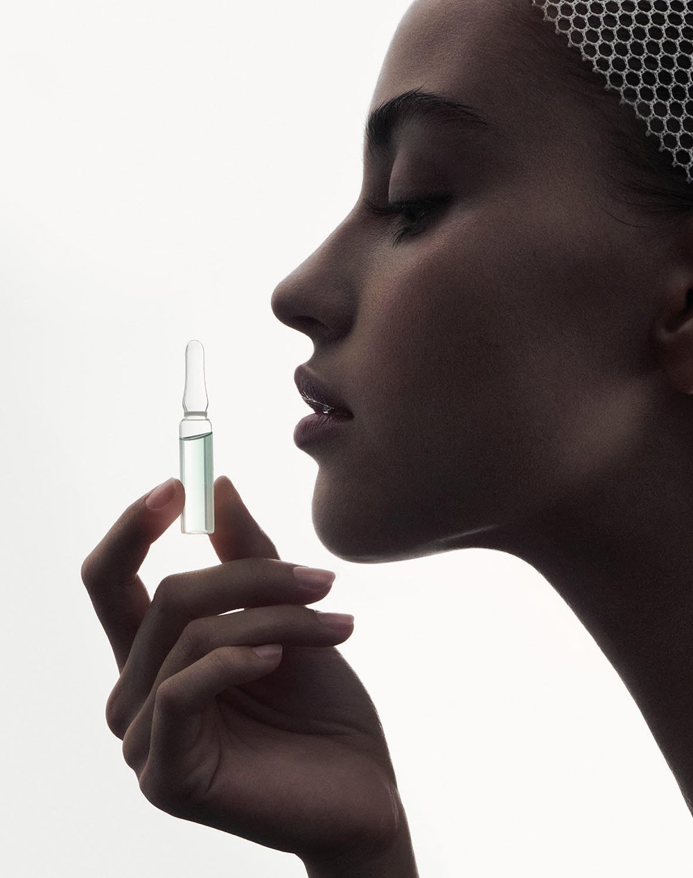 Close-up of a fair-skinned adult female in profile, holding a clear vial in her right hand, soft lighting against a white backdrop.