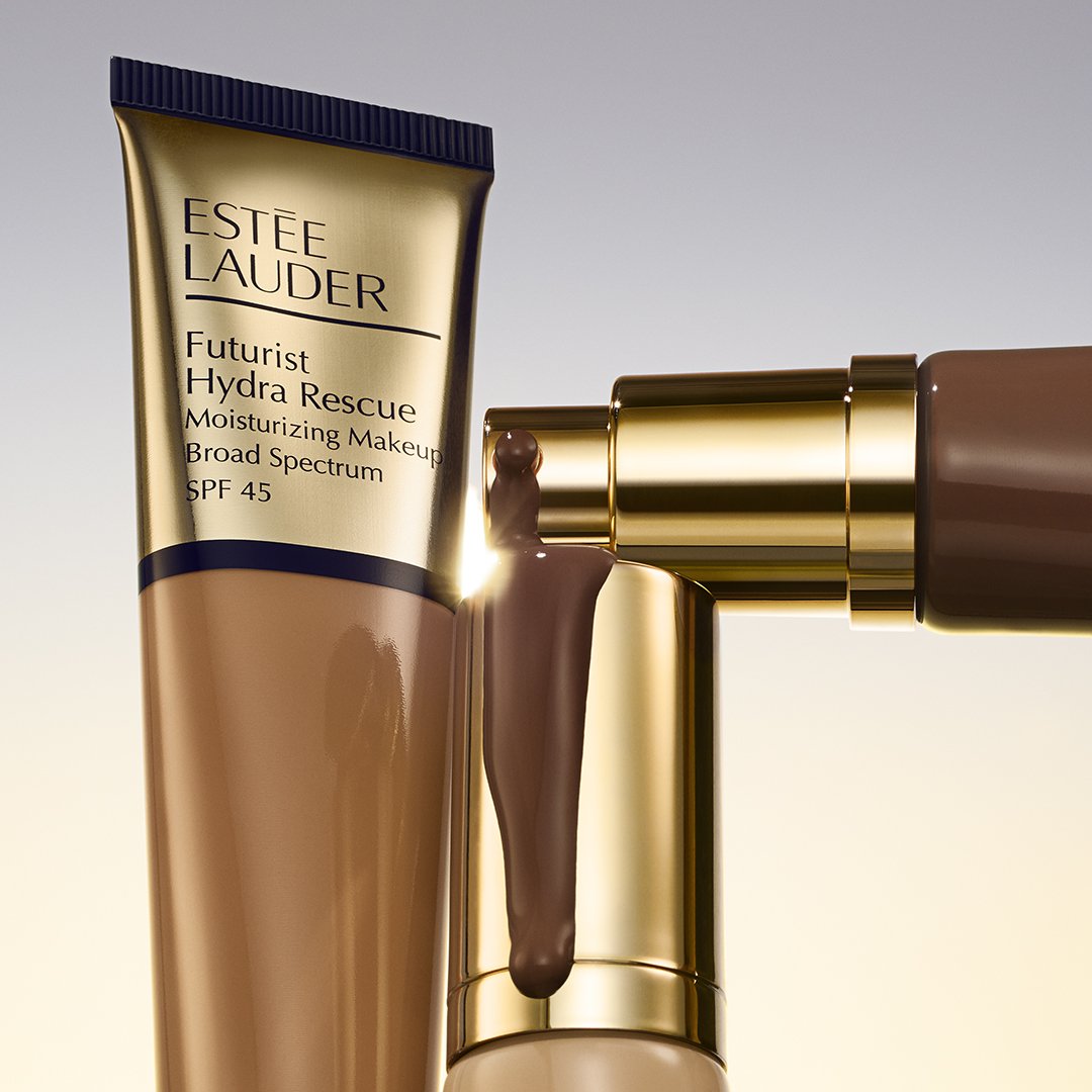 Close-up of Estée Lauder Futurist Hydra Rescue Moisturizing Makeup SPF 45 bottles and dripping foundation, golden accents, light background.