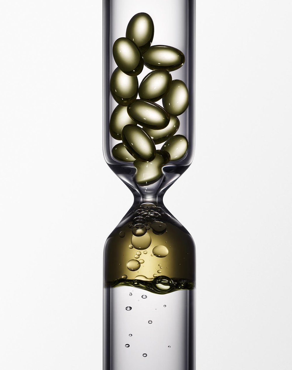 Hourglass-shaped glass container filled with golden capsules in the upper chamber, oil and bubbles in the lower chamber, against a white backdrop.